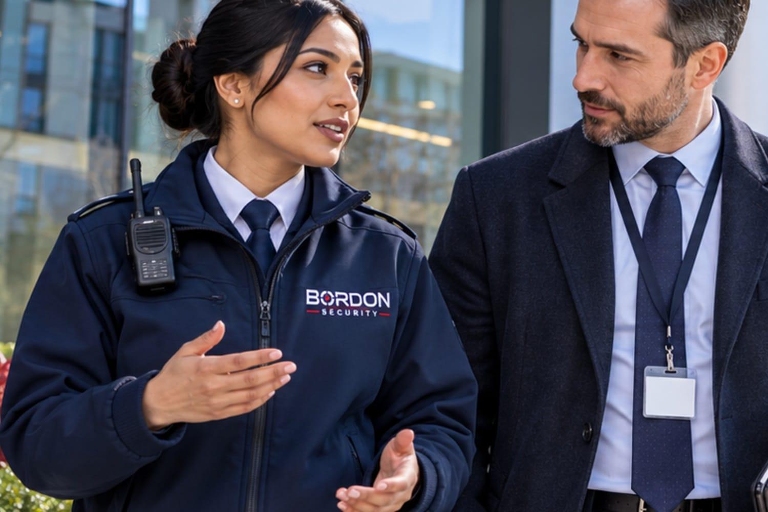 A Bordon Security officer in a navy jacket talks with a visitor wearing a lanyard outside a modern office building.